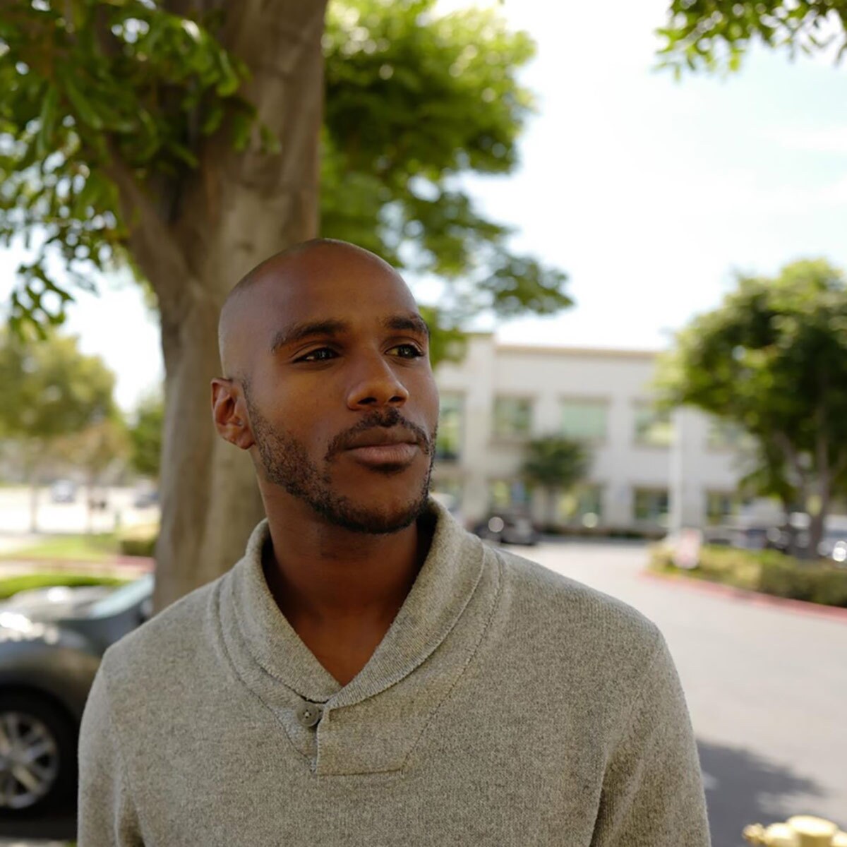 A young person wearing a beige sweater poses in front of a tree. 