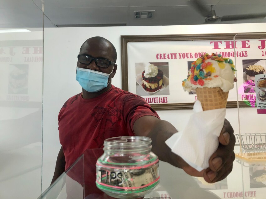 A man stands behind a corner and stretches his hand out, offering a scoop of ice cream on a waffle cone. The man is wearing a red t-shirt, a blue disposable face mask and black-rimmed glasses. On the counter in front of him is a tip jar filled with dollar bills. 