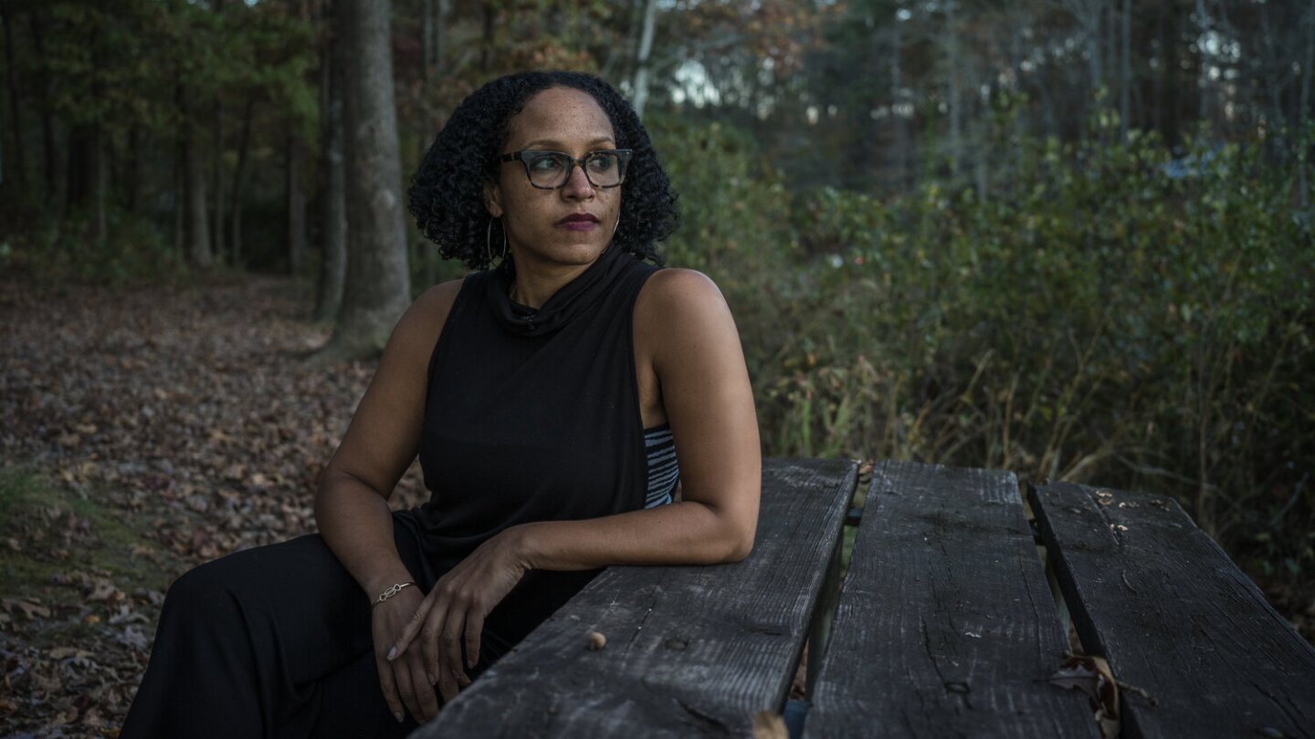 Gail sits at a wooden picnic table. She's looking off to the side. Behind her is lush, forest scenery.