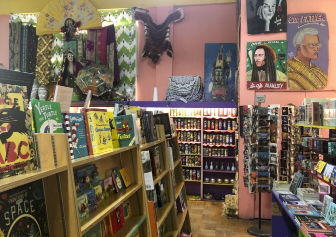 The interior at Los Feliz. The back end of the wall has a purple shelf displays rows of quirky Catholic candles. To the left of the photo is a shelf containing various books of different shapes, sizes and colors. To the right of the photo is a table containing even more books. A painting of Bob Marley hangs on the vibrant pink walls.