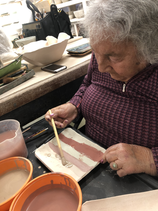 An elder woman paints on a piece of ceramics.
