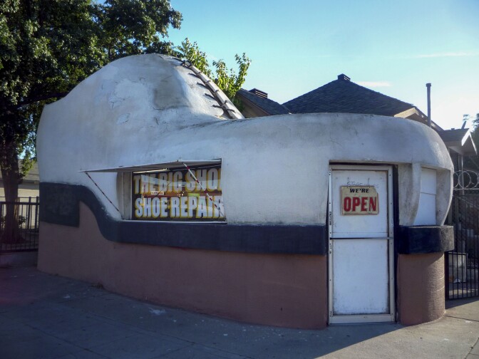 A giant shoe-shaped building stands with a white door with a sign reading, "We're Open." To the side of the building is a window with lettering that reads, "The Big Shop Shoe Repair." The shoe is predominantly white with a black sole and a reddish-brown base. The building looks worn, with parts of the white shoe dirty and flaking off.