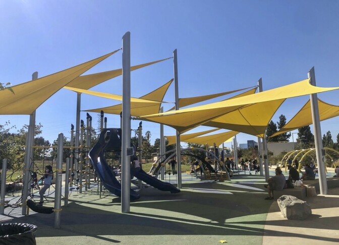 A playground with yellow tarp above providing shade. Children climb the various parts of the playground while parents watch from nearby benches. Above the playground is a clear bright blue sky.