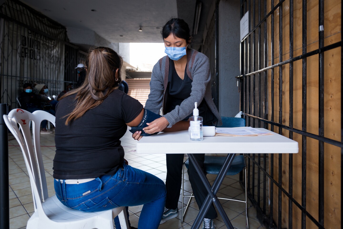 A woman in black shirt sits at a foldable table outside as she gets her blood pressure taken by a volunteer in a blue medical mask.