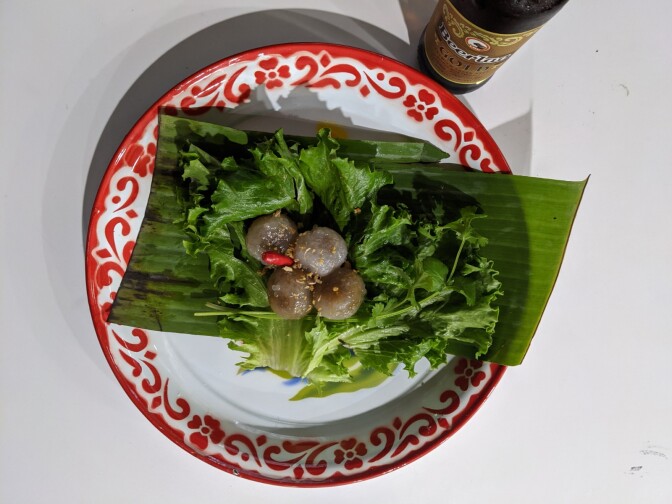 A plate of sakoo yat sai, tapioca dumplings filled with a savory filling and served with fresh vegetables. The dumplings are a light brown color and in the shape of four balls in the center of the plate. They sit on a bed of green lettuce and herbs.