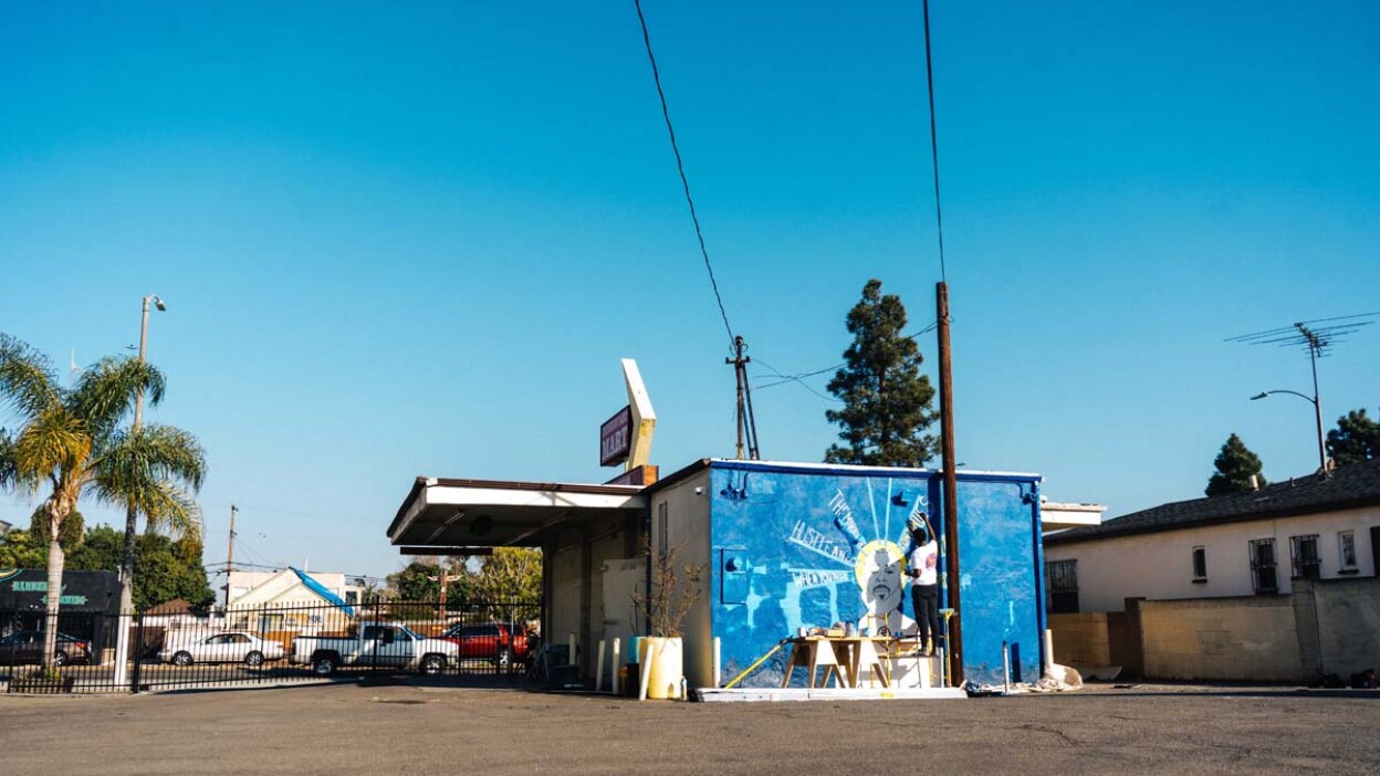 A wide shot of the Crenshaw Dairy Mart showing artist Oto-Abasi Attah painting a mural of Nipsey Hustle, "Saint Nip" | Courtesy of Crenshaw Dairy Mart