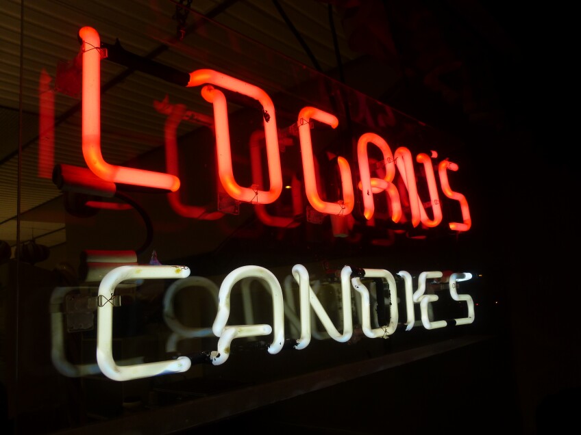A close up of a neon sign illuminating a dark environment. The neon sign reads, "Logan's Candies" in a rounded capitalized lettering. "Logan's" is in red and "Candies" is in white. 