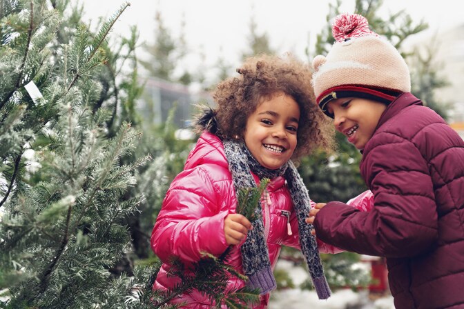 Two smiling children in puffy, colorful coats stand near snowy pine trees. istock