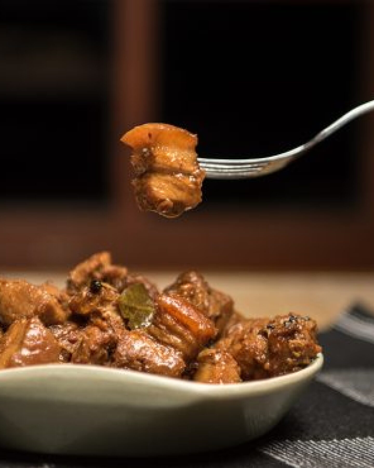 A hand raises a piece of meat from an adobo dish with a fork.