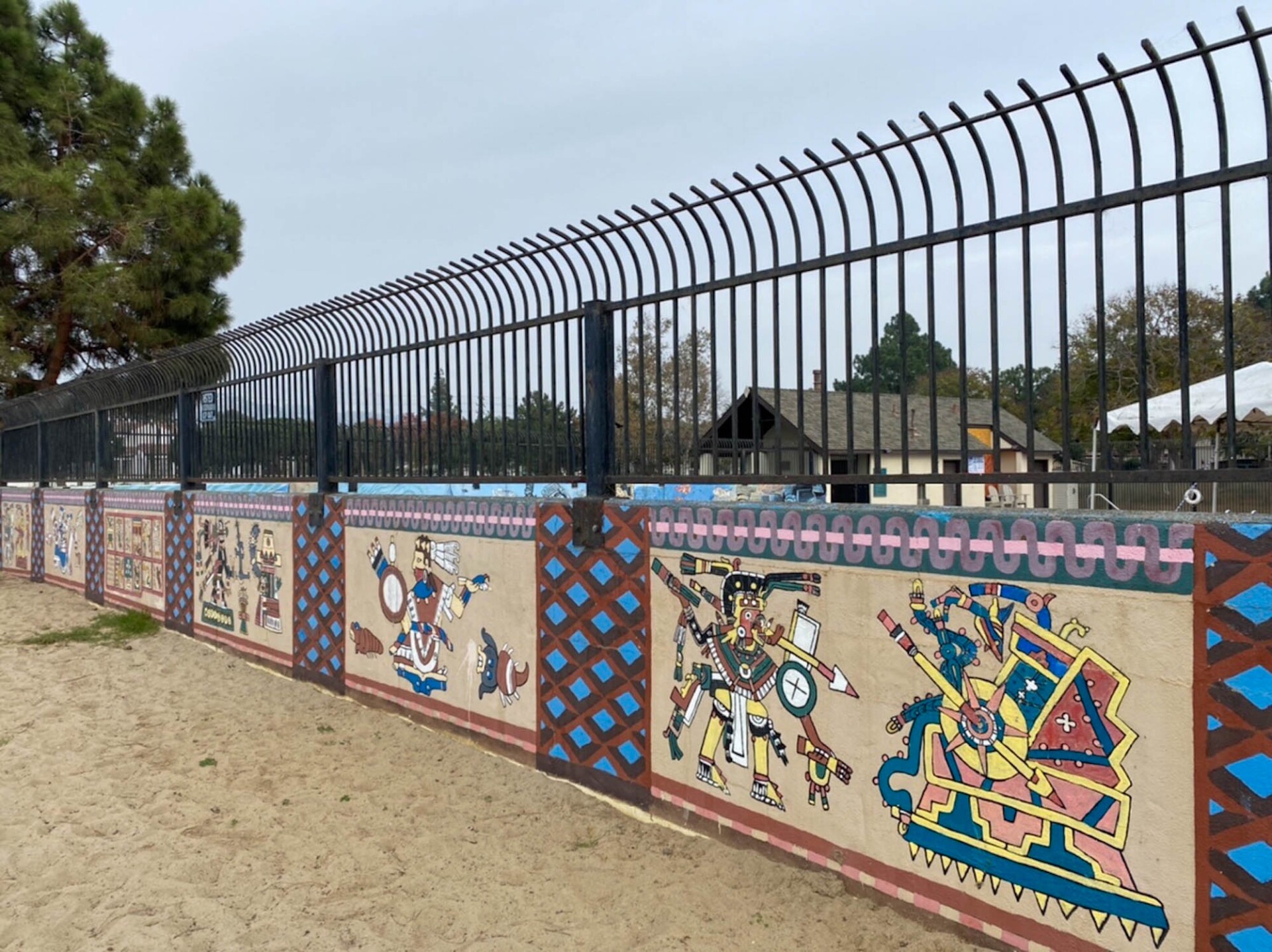 A short wall topped with black steel bar fencing features an intricate mural piece. The top and bottom of the wall is lined with intricate patterned borders. Every few feet, panels are divided by a criss-cross patterned border. In each panel are Indigenous imagery.