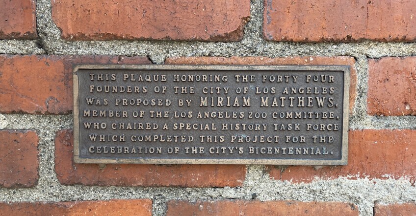 A metal plaque commemorating and acknowledging the efforts of Miriam Matthews is fixed onto a red brick wall. The plaque reads: "This plaque honoring the forty four founders of the city of Los Angeles was proposed by Miriam Matthews, member of the Los Angeles Zoo Committee, who chaired a special history task force which completed this project for the celebration of the city's bicentennial."