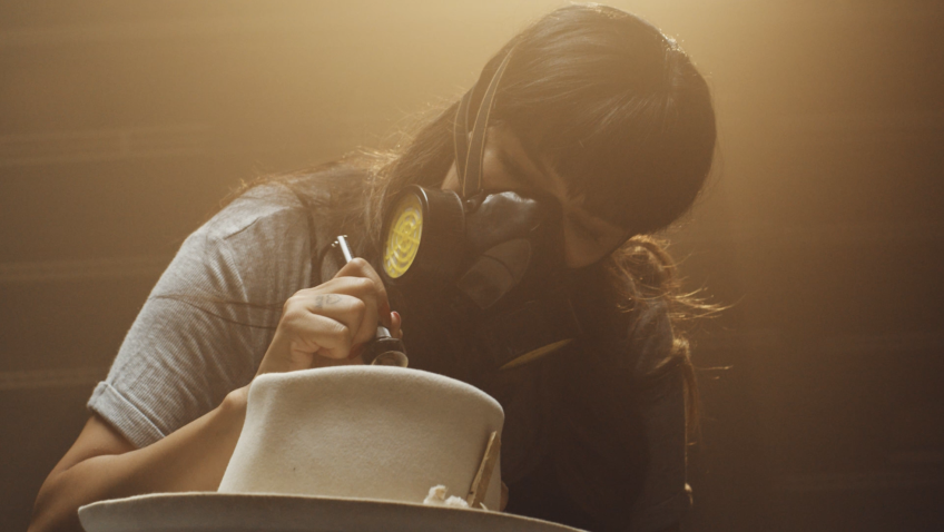 Pachuco Supply hatmaker Cinthya Marquez brands a hat with a branding pen. She is wearing a protective mask over her face as she leans over to work on the hat.