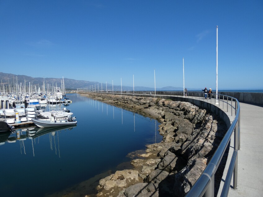 Boats are docked in still water protected by a rock wall where pedestrians walk atop of. Beyond, the mountains meet the sea against a bright, clear blue sky. 