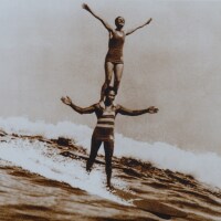 A sepia-toned photo of Duke Kahanamoku surfing a wave while Viola Hartman stands on his shoulders. The two have their arms outstretched as they ride the wave.