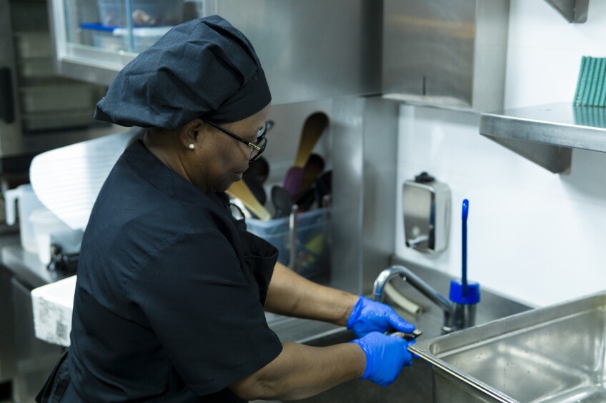A Black woman wearing blue protective gloves works at a commercial kitchen sink