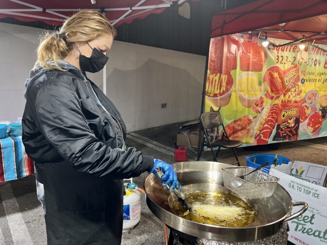 Minerva Osorno fries a quesadilla in a large pot of bubbling oil. She's frying the quesadilla outdoors under a tent. Minerva is wearing a black jacket, apron and surgical face mask. She's using a pair of tongs to flip the quesadilla.