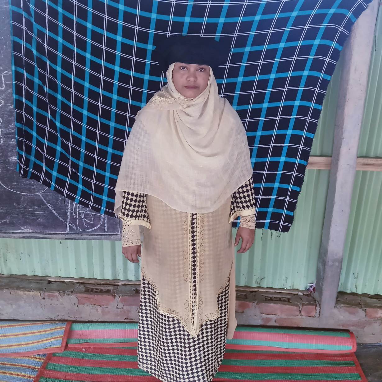 A woman in hijab, a black hat and and loose clothes stands in front of the camera.