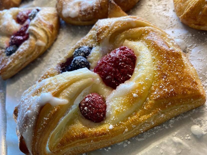 A close-up of a cheese danish with raspberries and blueberries on top. The baked good is dusted with white powdered sugar.