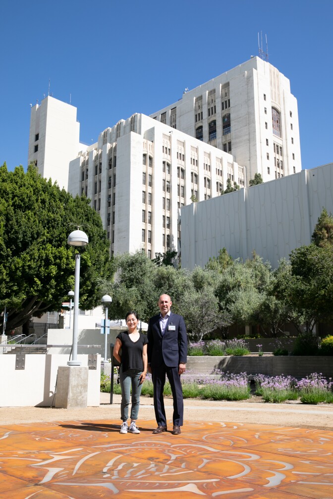 Phung Huynh, left, stands next to Jorge Orozco, right. They're both standing on the "Sobrevivir" art installation, a metal sculpture piece on the ground with floral etchings. Behind the duo, the LAC + USC Medical Center stands tall.