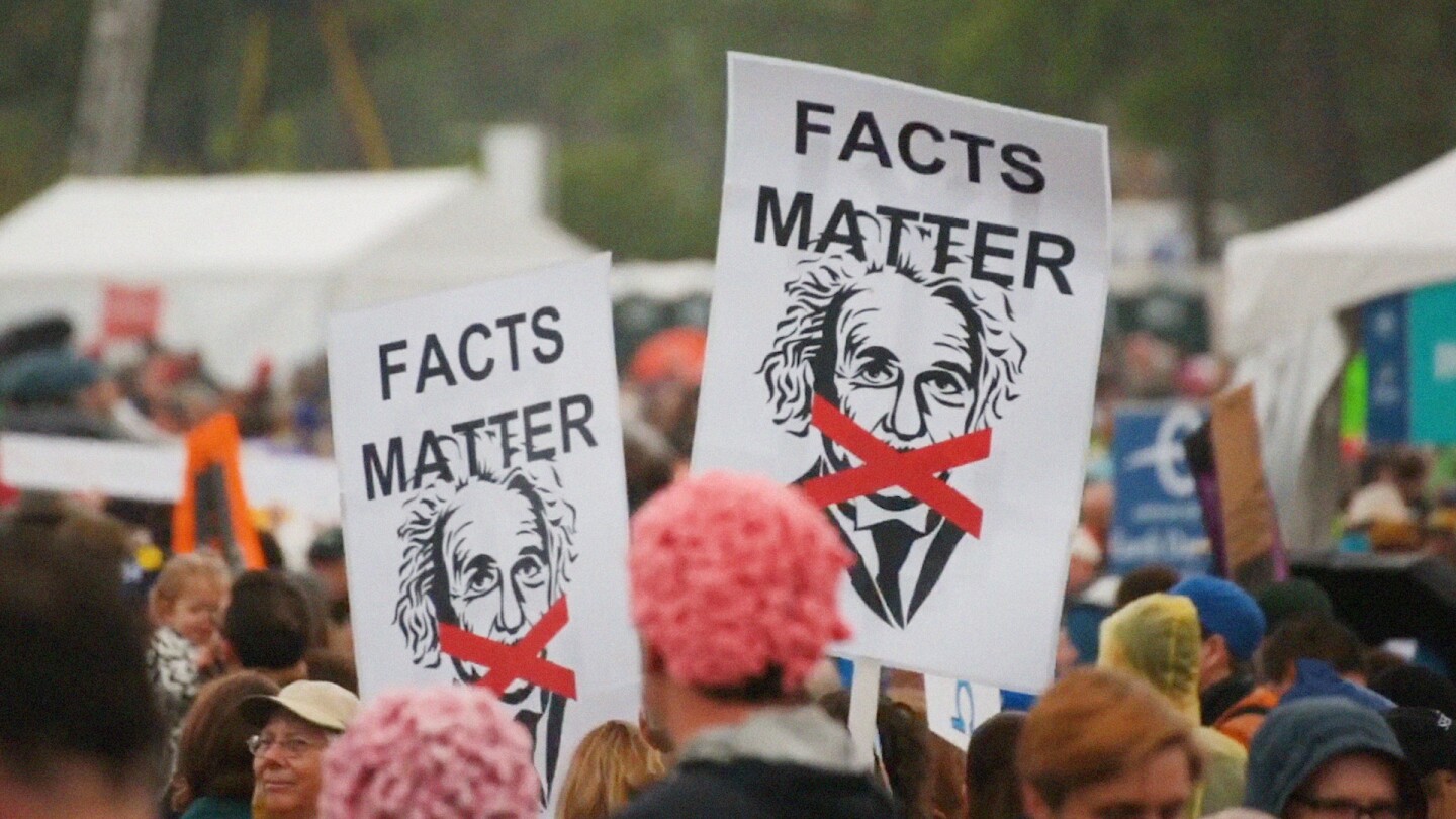 Protesters hold up signs depicting Albert Einstein and an "X" across his mouth, with the words "Facts Matter."