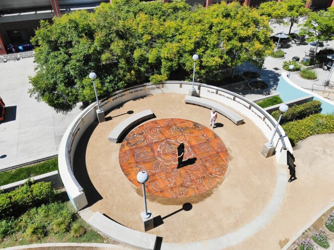 An overhead shot of Phung Huynh's circular sculptural piece "Sobrevivir." A woman stands in the middle of the piece and looks down at the etchings on the sculpture. Another woman stands to the side and looks at the artwork from a seating area.