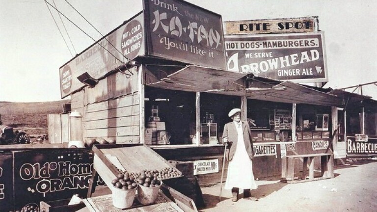 A sepia-tone historic photo of a man holding a cane standing in front of a food stand, surrounded by various crates, boxes, and advertising signs promoting cigarettes, candies, barbeque and more.