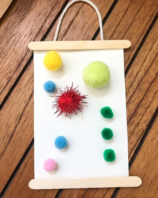 A hanging craft made out of popsicle sticks and colorful pom poms pasted onto a piece of paper with a string on top of it as a handle.