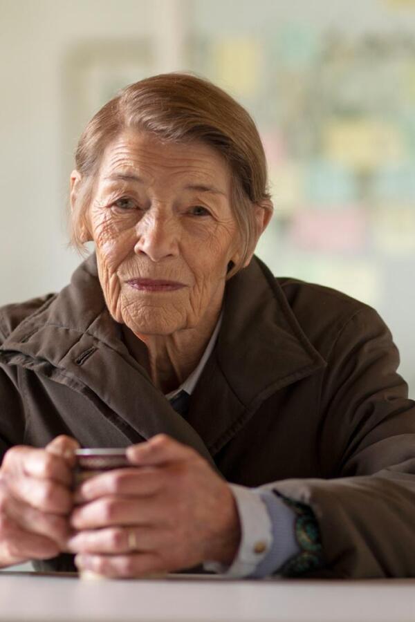 An elderly woman sits with mug in hand at a dining table.