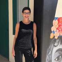 Annie Buckley is wearing an embroidered patterned black sleeveless top and black slacks. Her hair is a short, cropped cut. She's wearing black framed glasses and hoop earrings. She's learning against a black wall with a painted mural on it.