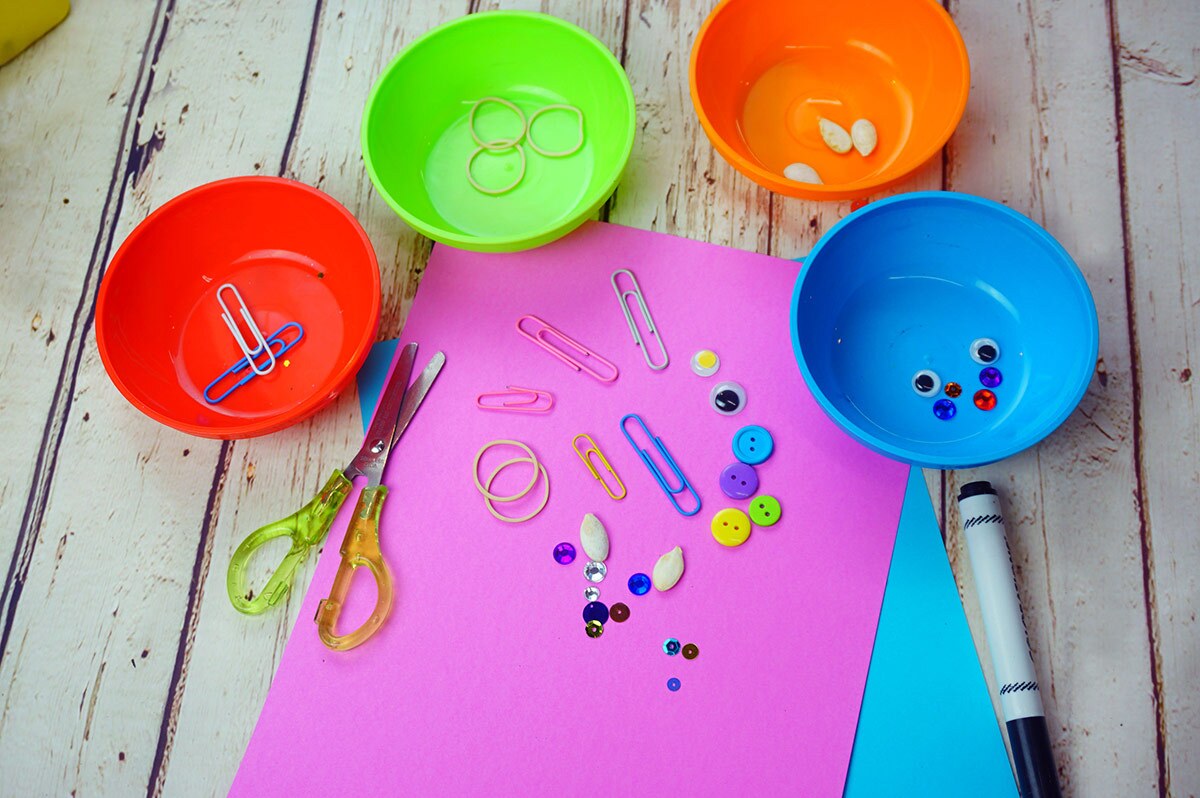 A piece of pink paper covered in small objects including sequins, paper clips and more surrounded by 4 colorful bowls filled with more of each of the items.
