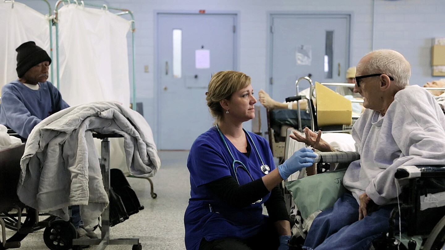 Nurse Tonia kneels to a patient in a wheelchair. | Still from "American Nurse" for "Link Voices"