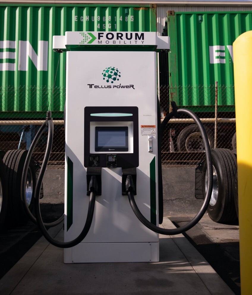 Green shipping containers are seen behind a white electric charging station that reads "Forum Mobility" and "Tellus Power" above a display screen and two charging cords