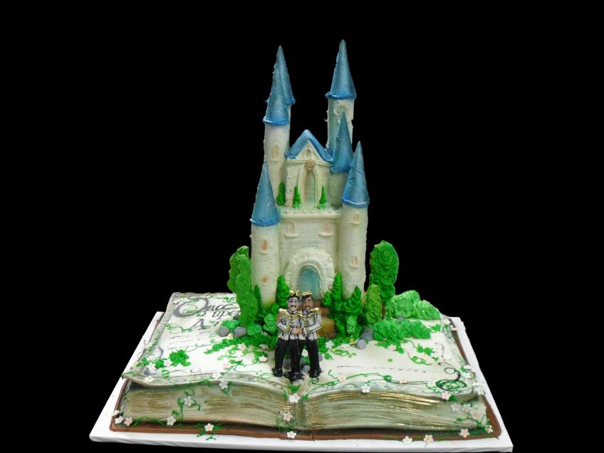 A wedding anniversary cake is shaped like an open storybook. In the center, a white castle with blue roofing juts out. In front of the castle, a fondant sculpture of two men dressed like kings or princes hug each other.