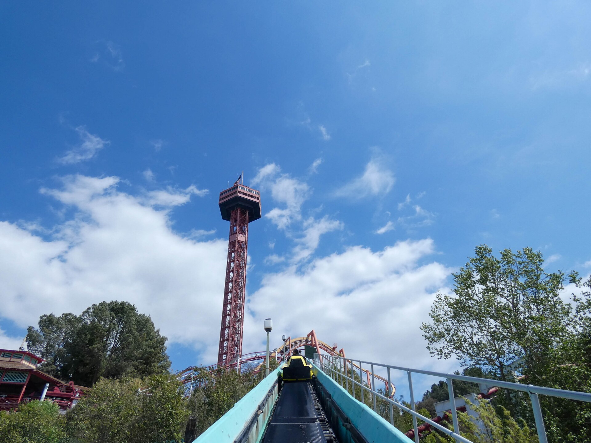 A point of view shot of a ride car being transported up a ramp via conveyer belt. Both sides of the ramp are enclosed by aqua blue bumpers. Beyond the ramp are parts of another ride — coaster tracks looping around. Beyond that in the near distance is a red view tower.