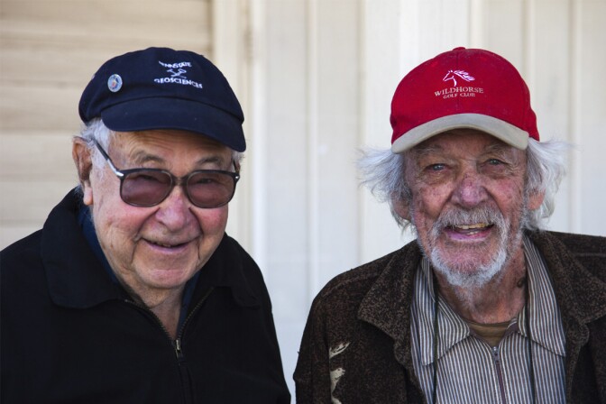 Two elderly men sit next to one another and look direct to camera. The man on the left is wearing a dark navy blue cap with "Penn Sttate Geosciences" embroidered on the front and a pin on the side. The man on the right is wearing a red cap with "Whitehorse Golf Club" embroidered in white on the front of his cap.