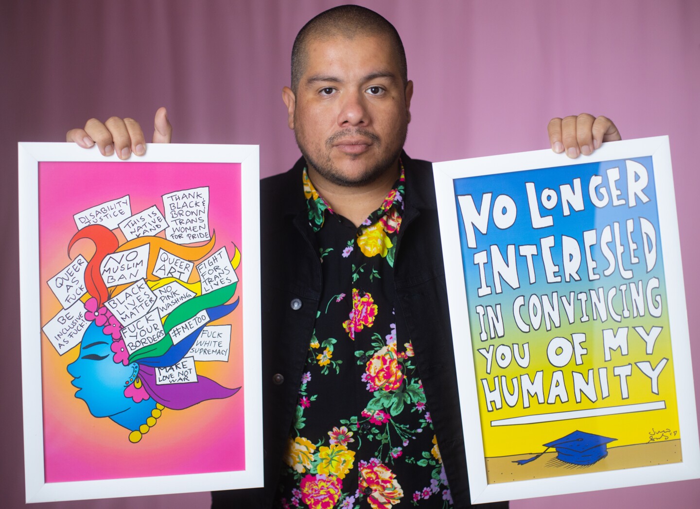 Julio Salgado is wearing a floral print shirt and a black jacket while holding up two pieces of his art on each hand. The artwork on his left features the side profile of a woman with multicolored hair and statements like, "Black Lives Matter," "#MeToo," "Make Love Not War," and "Thank Black and Brown Trans Women for Pride." The artwork on the right reads, "No Longer Interested in Convincing You of My Humanity," with a graduation cap at the bottom. Salgado is standing in front of a pink background.