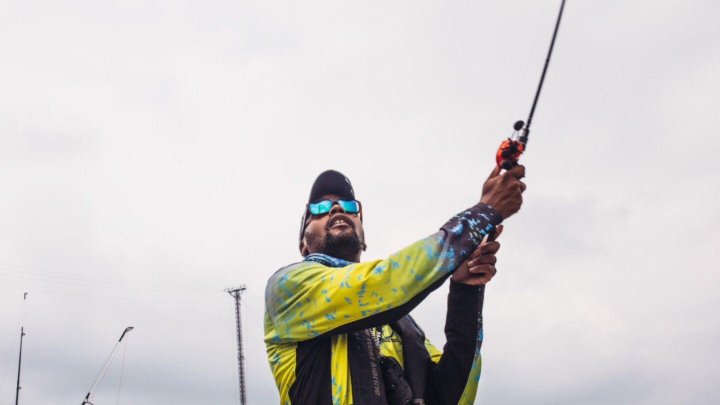 A man casts a fishing line. Behind him is a gloomy, gray-blue sky. The man is wearing sunglasses with teal blue lenses and a bright yellow jacket.