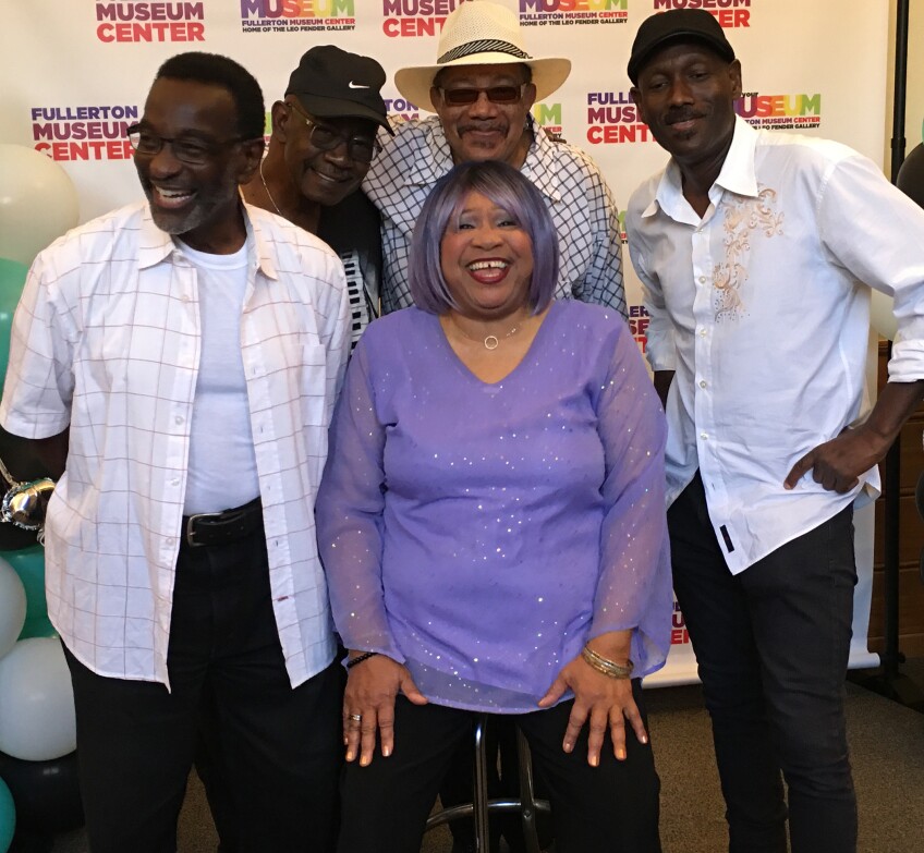 Members of Sista Jean's Blues Machine stand in front of a wall that reads, "Fullerton Music Center." Sista Jean sits in the middle, wearing a sparkling purple top with matching purple streaks in her hair. Around her, four men huddle and smile for the photo.