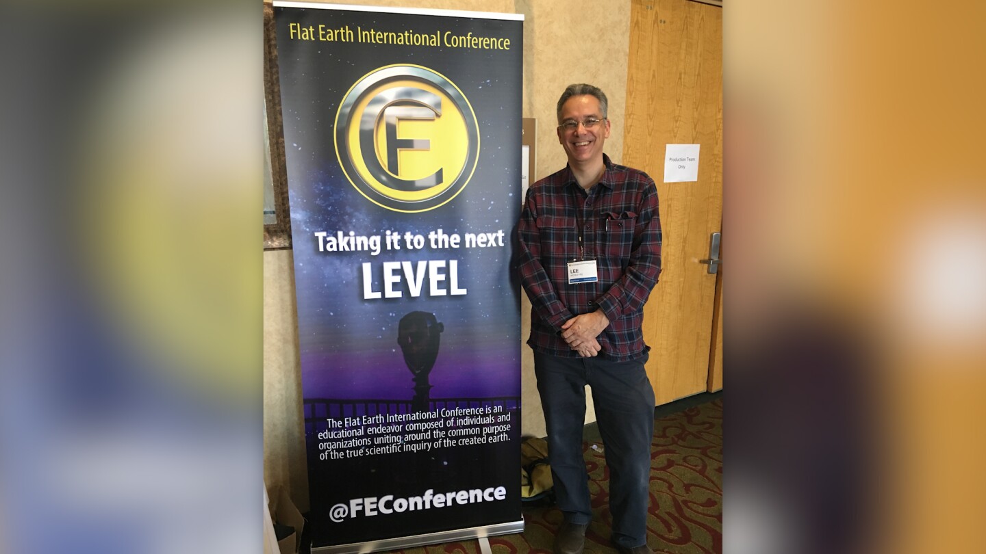 Lee McIntyre poses next to a poster promoting a flat earth conference.
