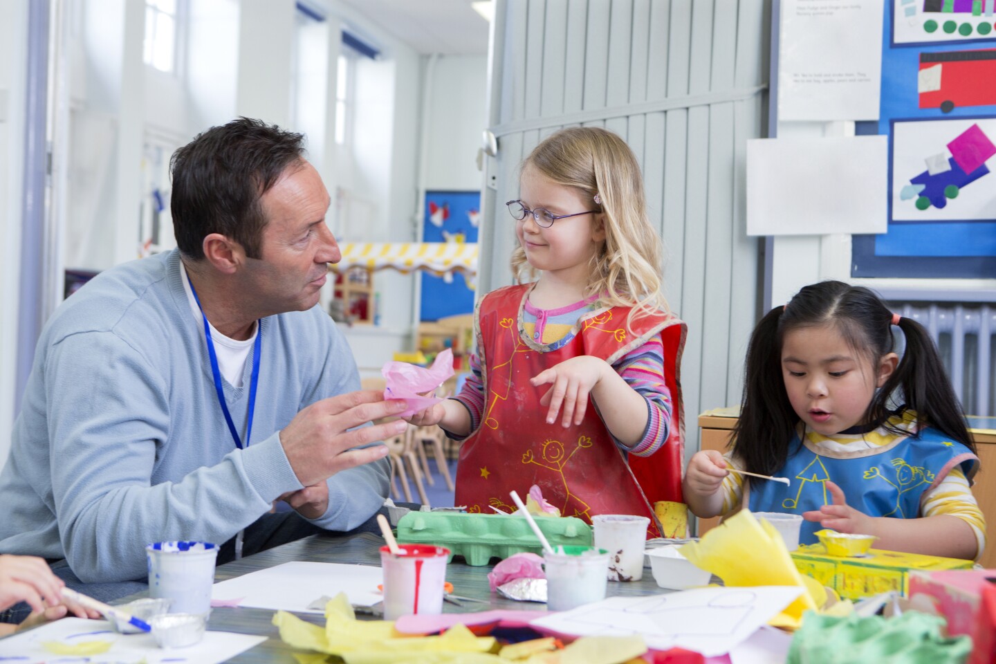 Male teacher talking to one of his nursery students in the classroom. They are doing arts and crafts.