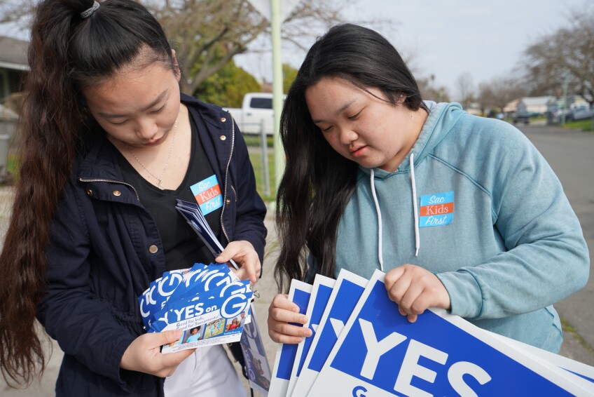 Two Asian American youth hold up signs and flyers saying "Yes on G"