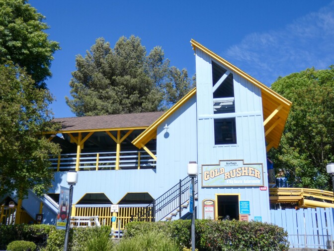 A light blue angular building made out of vertical wood paneling and brown tiled roofs with a bright yellow under-paneling. A sign on the face of the building reads, "Gold Rusher: A Rip Roarin' Rollercoaster." The building is asymmetrical with both sides of the roof coming at different angles.