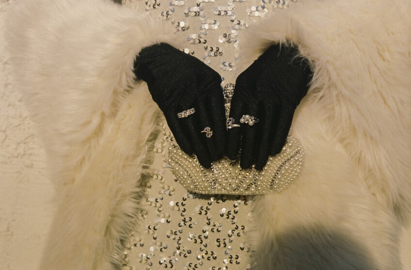 A close-up of Muxxxe's hands, covered with black gloves, delicately holding a bedazzled pearl clutch. Bedazzled silver rings decorate their fingers. Their sparkly white dress and faux fur coat create a texture background behind their hands.