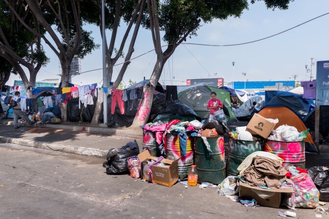 Barrels of trash line a road. Behind it are various tents. Clothes hang to dry between sparse trees by the tents.