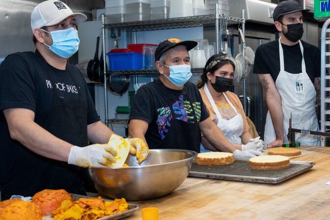 Four workers wearing surgical face masks and aprons stand behind a wooden counter and look intently at something out of frame. In front of them are metal trays and bowls. The metal tray has two layers of white round cakes sitting on it while the bowl has pureed fresh pumpkin.