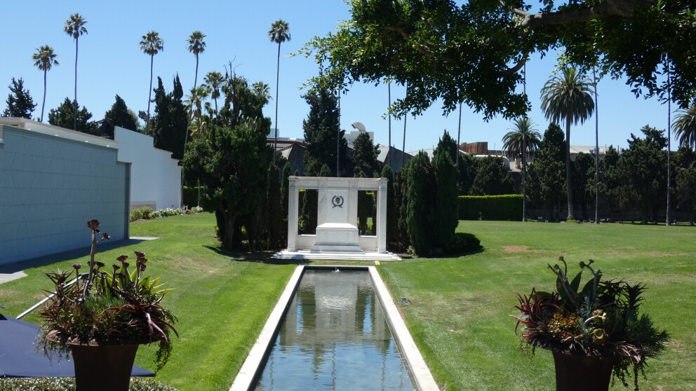 A reflection pool leads up to a white marble structure standing in the middle of a green field. Tall palm trees stand beyond. 