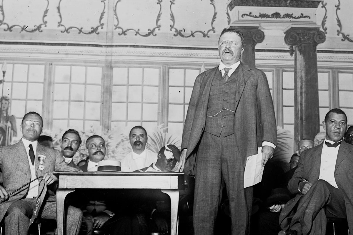 Theodore Roosevelt (foreground) stands on stage before an audience with Booker T. Washington sitting behind him. | Still from "City Rising: The Informal Economy"