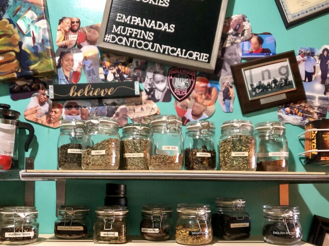 Looseleaf tea in large glass jars are arranged on two shelves. Behind it is a bright teal wall covered in various wall decor and personal photos, arranged in a collage style.