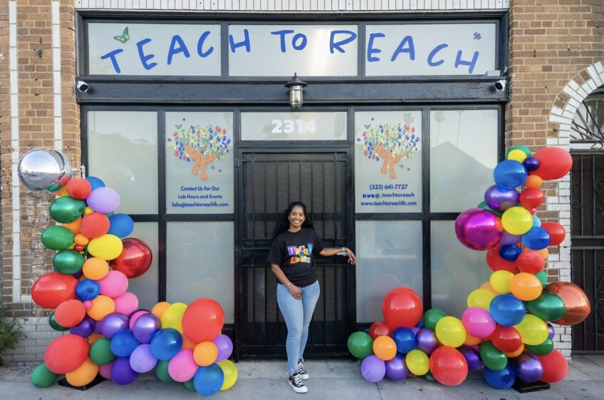 A young woman stands outside a storefront that says "Teach to Reach"