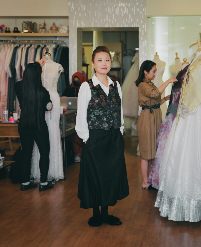 Laura, a hanbok maker, stands in her shop with dresses and tailors.